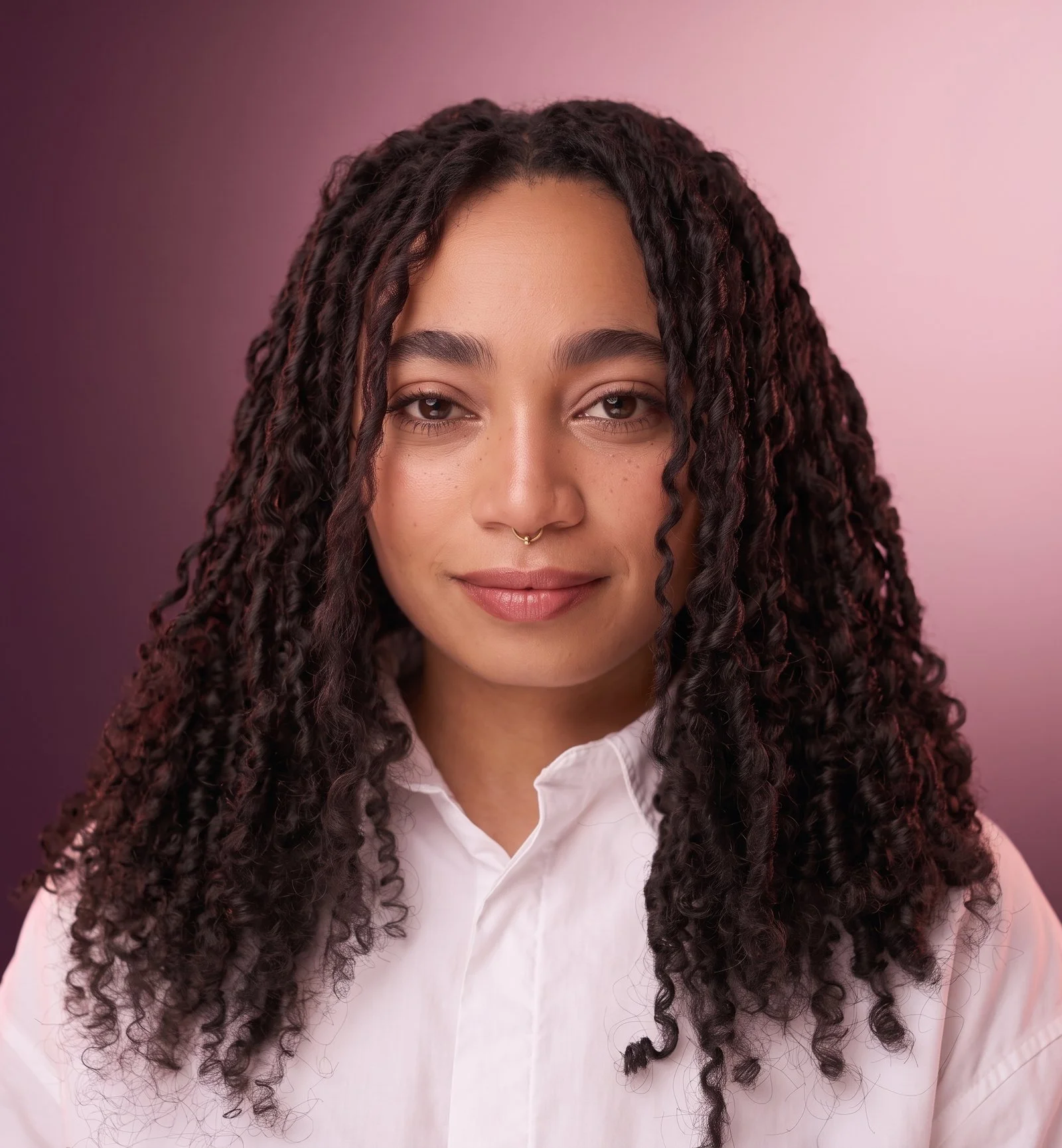 Mujer con cabello rizado largo y oscuro, camisa blanca y fondo rosa, mirando a la cámara.